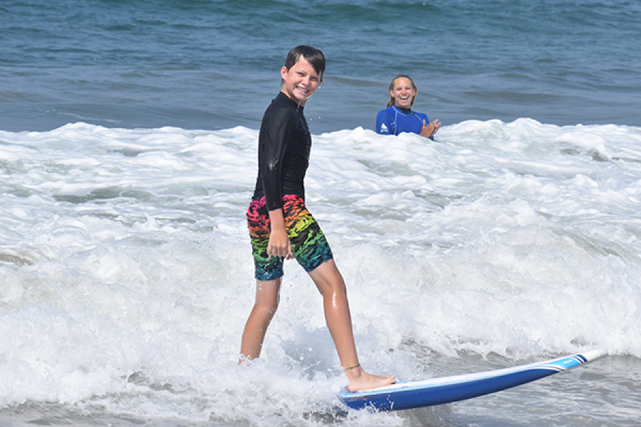 child on surf board smiling at camera with surf instructor in background cheering them on